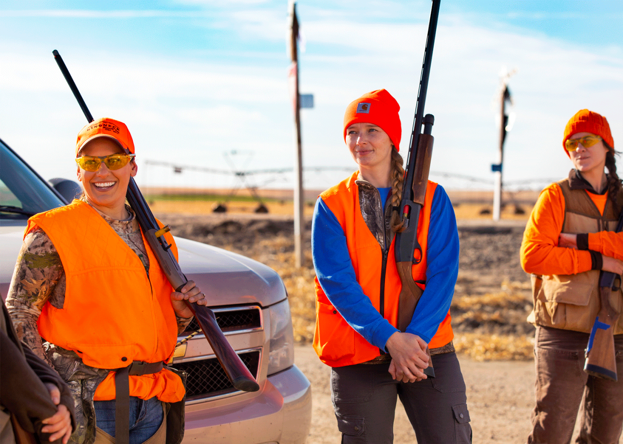 Three women wearing hunter orange vests and hats holding guns stand next to a pickup truck.