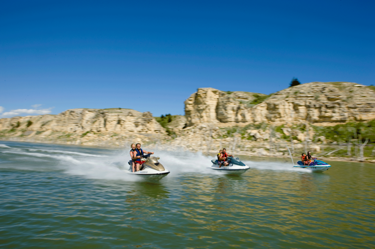 Three jet skis, each carrying two riders in life jackets, speed across a lake surrounded by tall rocky cliffs under a bright blue sky.
