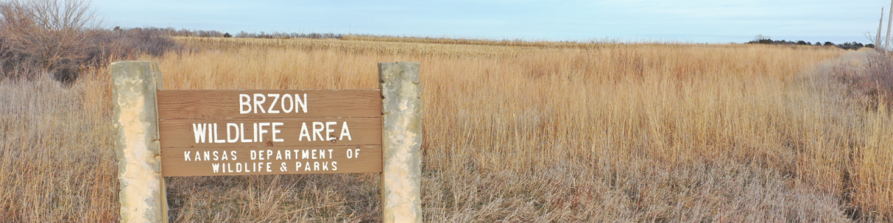 A wooden sign reads BRZON Wildlife Area Kansas Department of Wildlife and Parks. The sign stands among tall prairie grasses with open land stretching into the distance behind it.