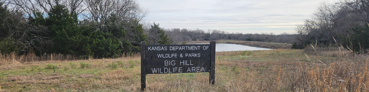 A wooden sign reads Kansas Department of Wildlife and Parks Big Hill Wildlife Area. The sign stands in a grassy clearing with a pond and tree line visible in the background under a cloudy sky.