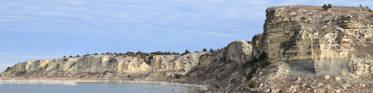 Layered rock cliffs rise above a body of water, showing distinct horizontal bands of stone. The rugged formations overlook the lake, with sparse vegetation scattered along the rocky slopes.