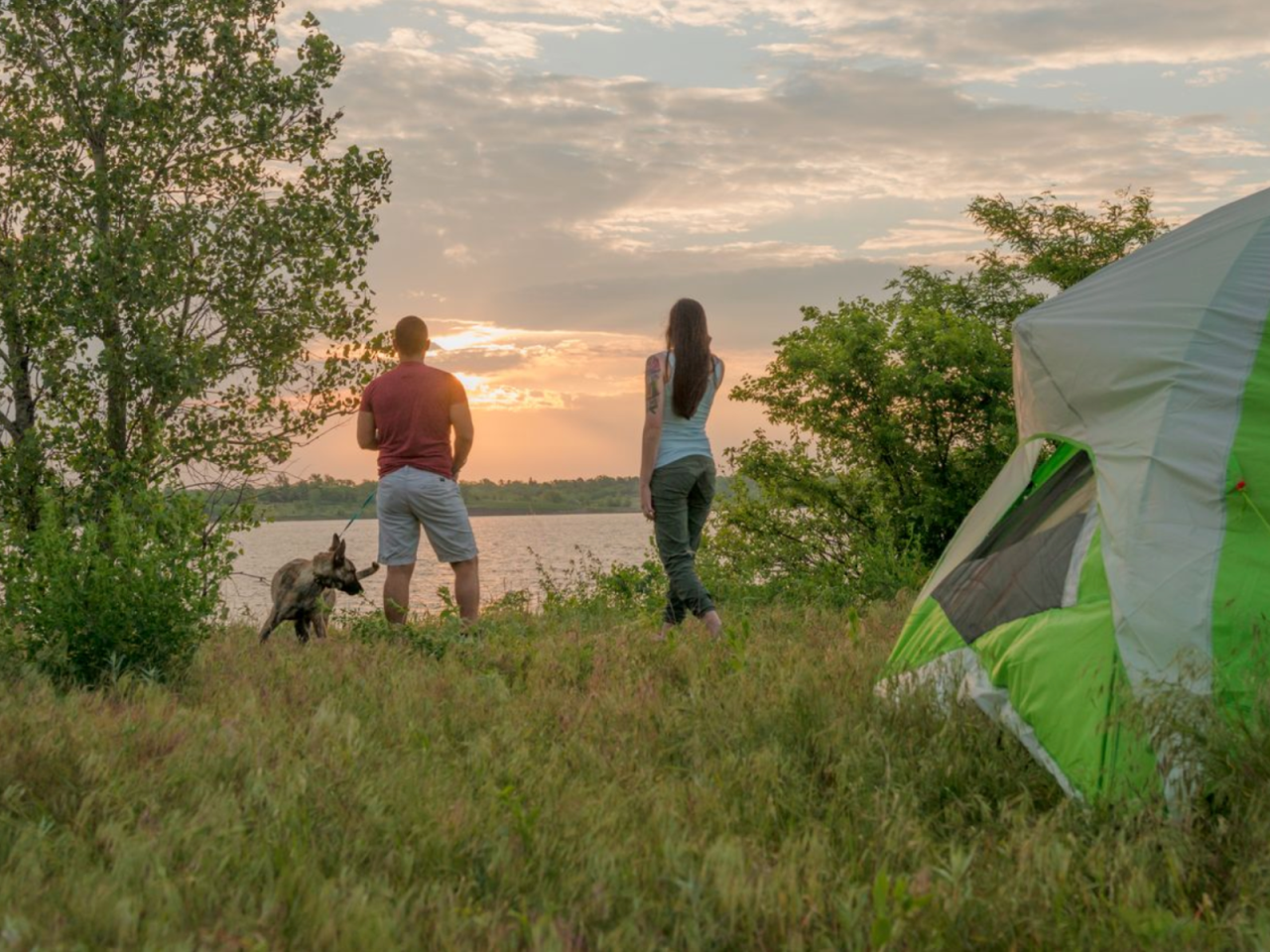 Two people and a dog by a lake at sunset, with a tent and trees nearby.