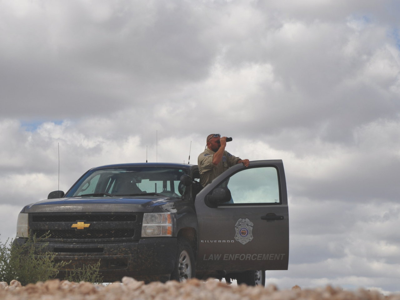 Officer with binoculars next to law enforcement vehicle.