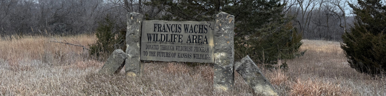 Stone entrance sign reading Francis Wachs Wildlife Area, donated through wildtrust program to the Future of Kansas Wildlife that stands between large rock posts, surrounded by tall dormant grasses and trees in a natural setting.