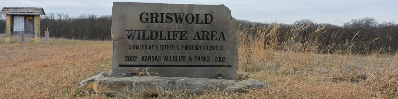 Stone sign reading "Griswold Wildlife Area- Donated by E.Ester & F.Arleigh Griswold- 2002 Kansas Wildlife & Parks 2002"  stands in a grassy field, with a small information kiosk and leafless trees visible in the background.