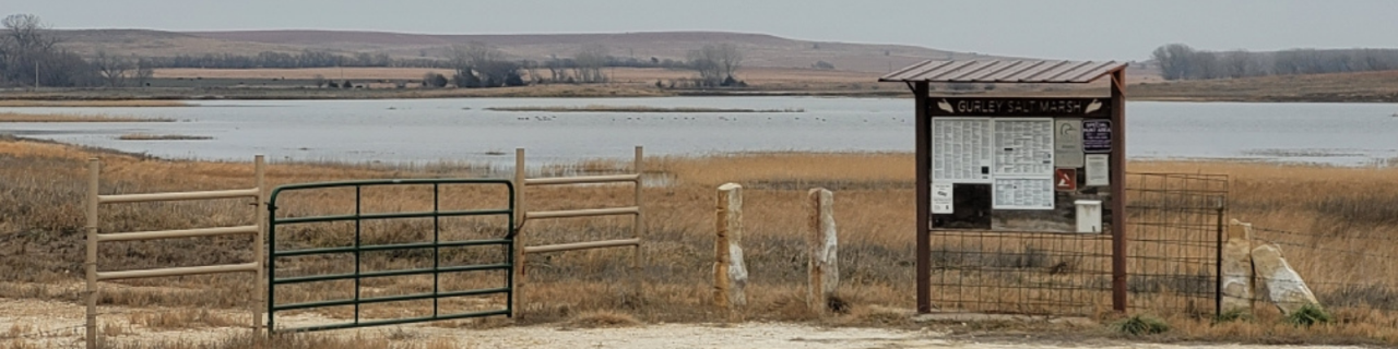 A gated entrance and informational sign overlook a large marsh. Water and low vegetation extend into the distance across a gently rolling landscape.