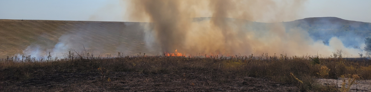Smoke rises from a controlled burn across a grassy field. Flames are visible near the ground, with hills in the background.