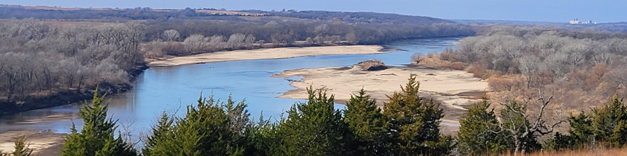 A river winds through a wide valley bordered by trees and sandbars. The view overlooks the water from an elevated position.
