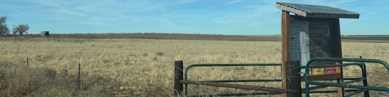 A gated entrance with an informational kiosk stands along a fence line. Beyond it, a wide, flat prairie stretches under a clear blue sky.