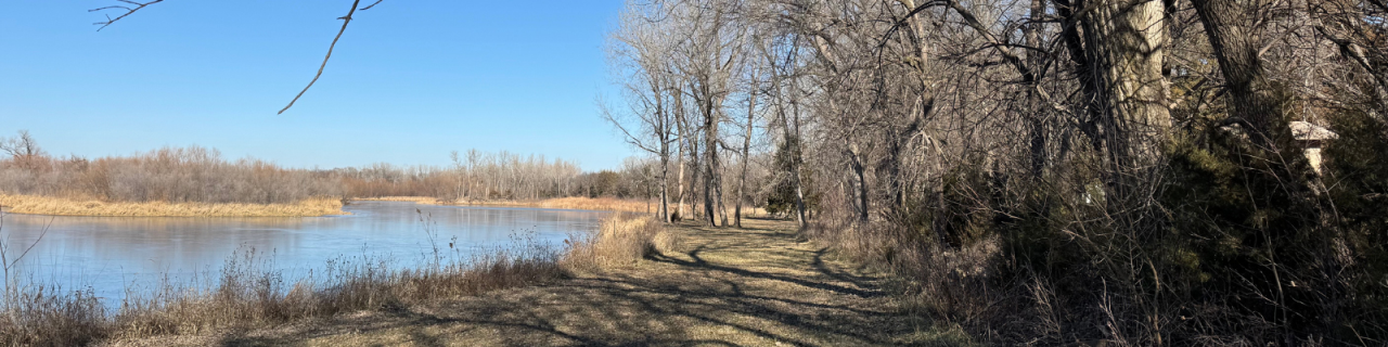 A calm river curves alongside a grassy path lined with leafless trees. The water reflects a clear blue sky.