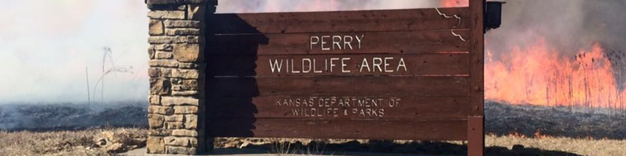 A wooden sign labeled "Perry Wildlife Area-Kansas Department of Wildlife & Parks"  stands in front of a prescribed burn, with flames and smoke rising behind it.