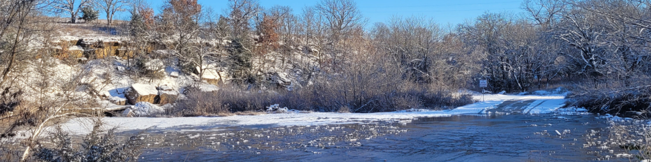 A shallow river flows through a rocky, tree-lined area with light snow covering the ground.