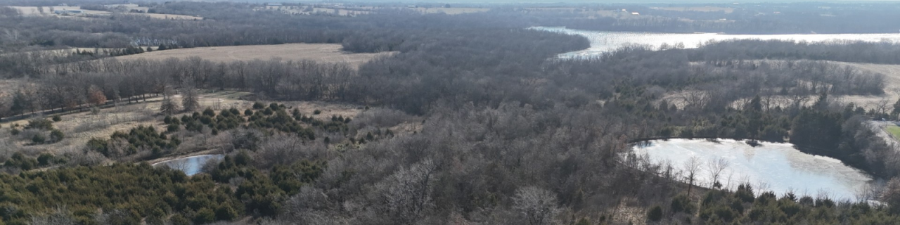 An aerial view shows a patchwork of trees, ponds, and open land with water visible in the distance.