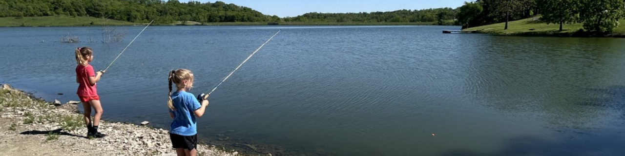 Two children stand on a rocky shoreline fishing in a calm lake surrounded by green trees and grassy hills.