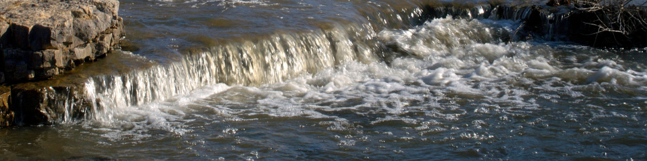 Water flows over a low rocky spillway, creating a small waterfall with rippling currents below.