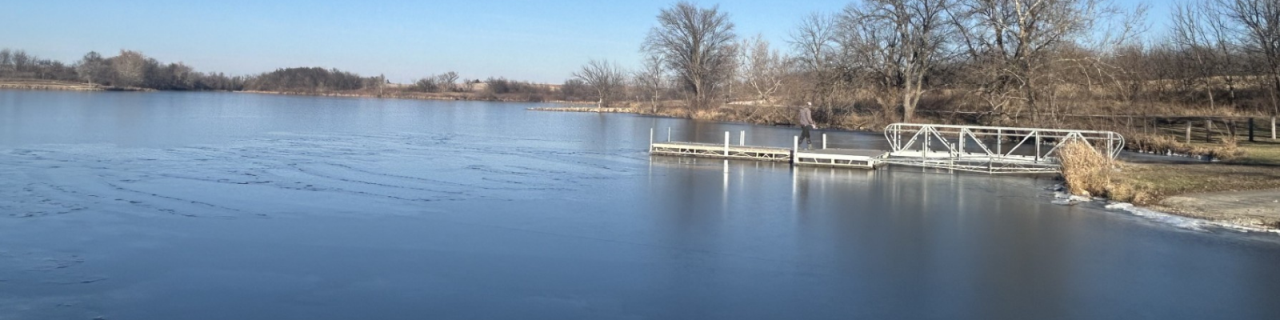 A lake with a small dock with a man walking on, extends into the water, surrounded by leafless trees and winter grass.