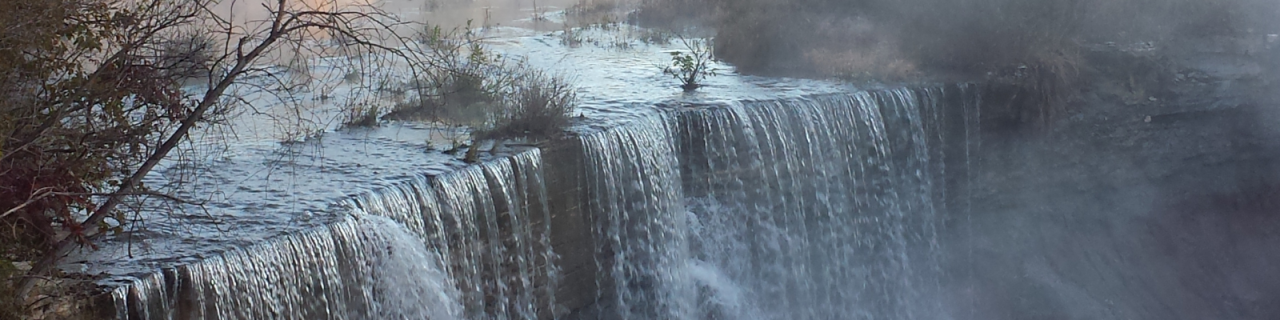 Water cascades over a larger dam, forming a wide waterfall surrounded by rocks and trees.