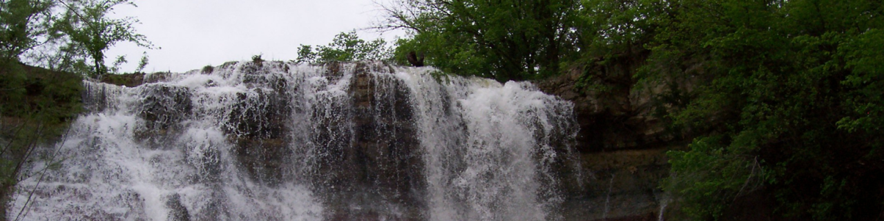 Water cascades over a larger dam, forming a wide waterfall surrounded by rocks and trees.