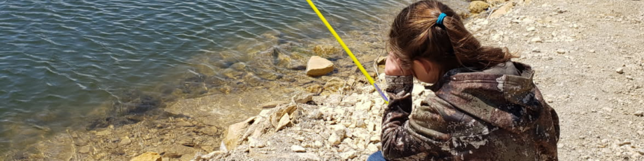 A young person sits on a rocky shoreline fishing, holding a rod over clear water with visible stones beneath the surface.