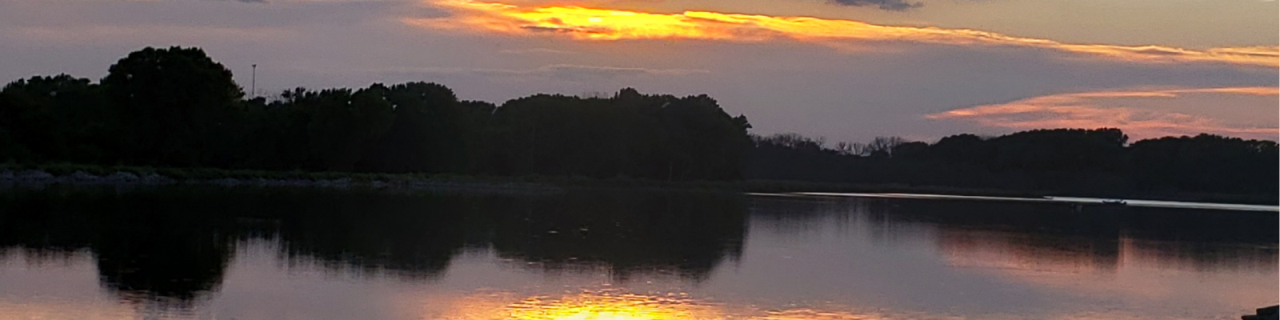 A lake at sunset reflects orange and pink hues from the sky, with dark tree silhouettes lining the far shoreline.