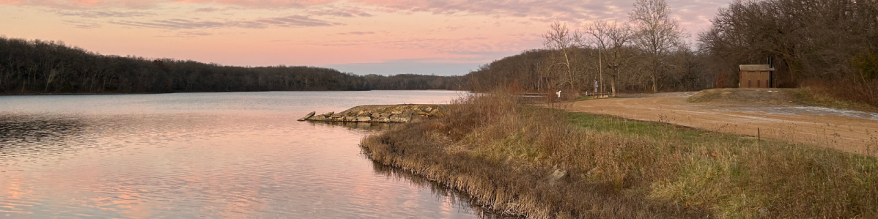 A peaceful lake at dusk with a small peninsula and nearby vault toilet sits beside a gravel road, surrounded by trees under a pastel-colored sky.