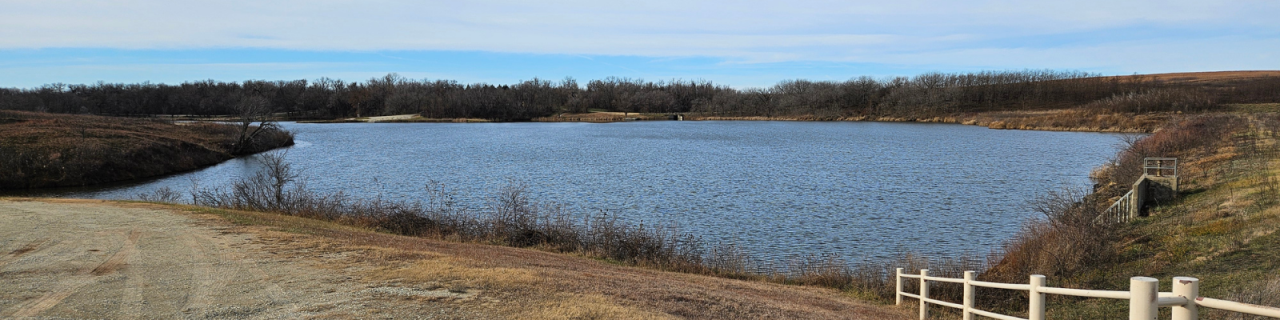A lake is surrounded by rolling prairie and leafless trees, with a metal fence and concrete structure to the right under an open sky.