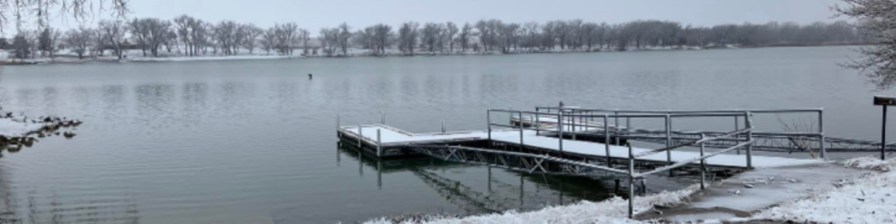 A snow-dusted dock extends into a still lake, with frost-covered banks and bare trees in a winter landscape.