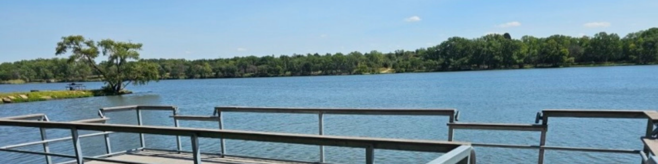 A metal fishing dock extends over a calm lake, with tree-lined shores in the distance.