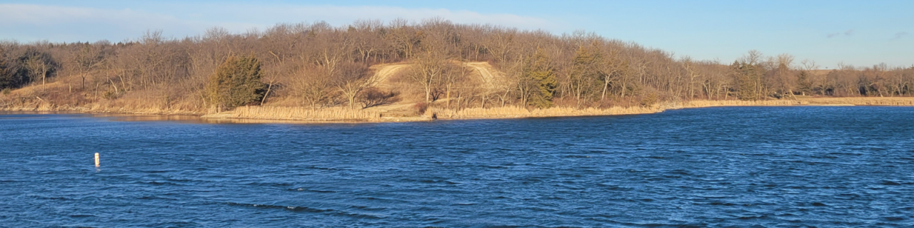 A blue lake ripples with a white floating bouy with a tree-covered hill rising along the far shoreline.