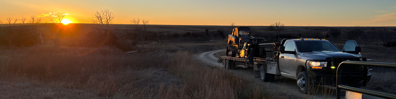 A pickup truck towing equipment drives along a dirt road beside open prairie at sunset.