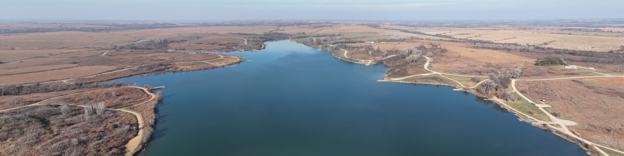 An aerial view shows a winding lake surrounded by open prairie, dirt roads, and scattered trees.