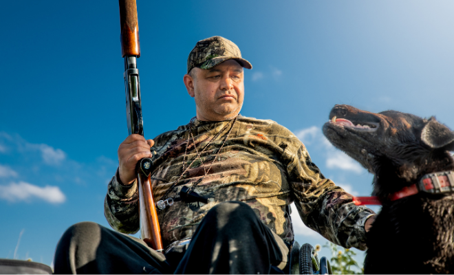 A man in a wheelchair holds a hunting rifle while sitting beside a hunting dog in an outdoor setting.