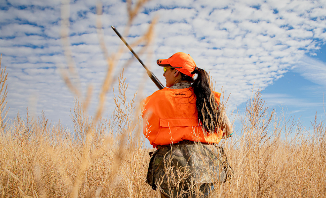 Person in bright orange hunting gear standing in a field under a cloudy sky.