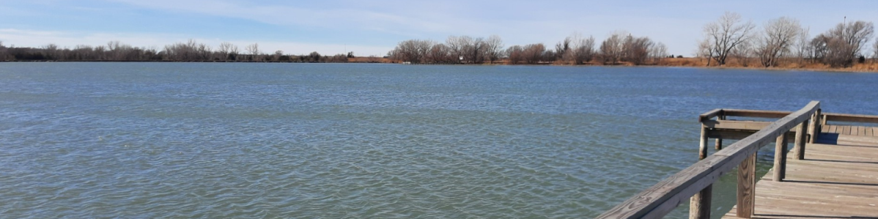 A wooden fishing dock extends over a calm lake with small ripples, while leafless trees line the opposite shoreline under a clear blue sky.