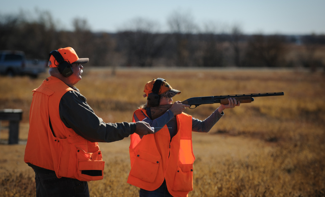 Two people in orange safety gear, with one aiming a shotgun outdoors.