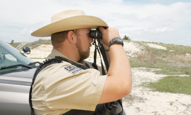 Man in a beige uniform and cowboy hat using binoculars outdoors near a vehicle.