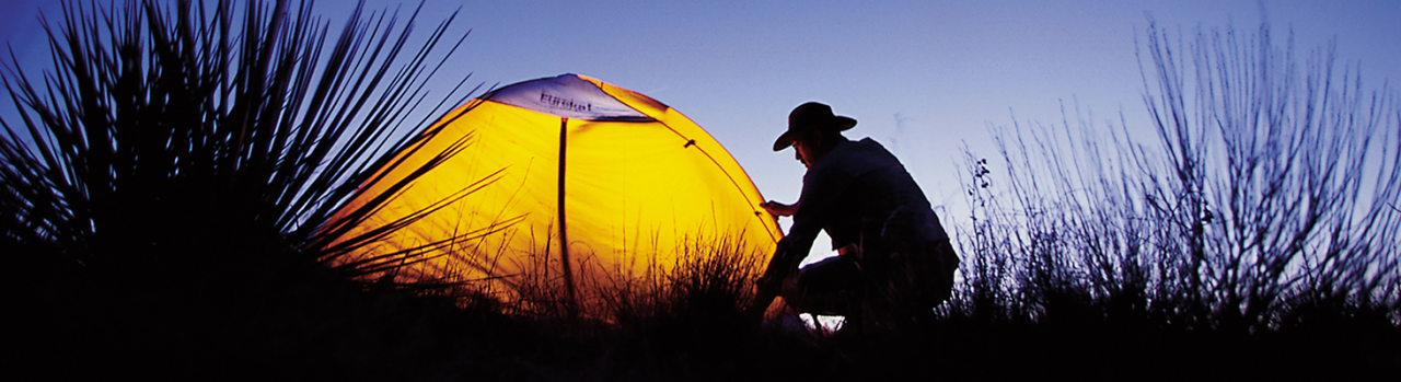 A person in a hat kneels to adjust a brightly lit yellow tent at dusk, surrounded by dark grasses and foliage against a twilight sky.